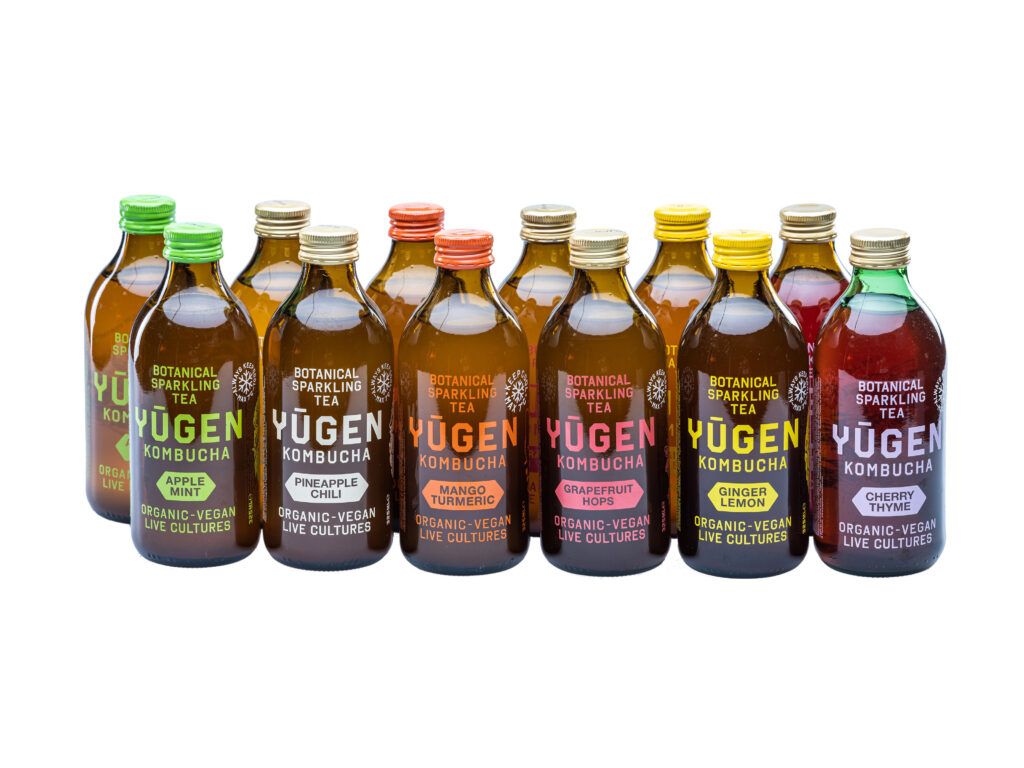 Row of Yugen Kombucha bottles with various flavors including Apple Mint, Pineapple Chili, Mango Turmeric, Grapefruit Hops, Ginger Lemon, and Cherry Thyme, labeled as organic and vegan with live cultures.