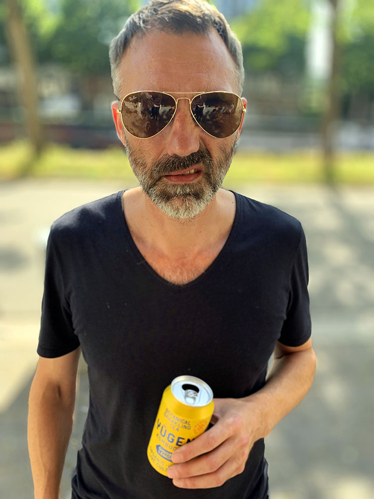 Homme en t-shirt noir et lunettes de soleil aviateur tenant une canette de Yugen Kombucha au gingembre et au citron à l'extérieur par une journée ensoleillée.